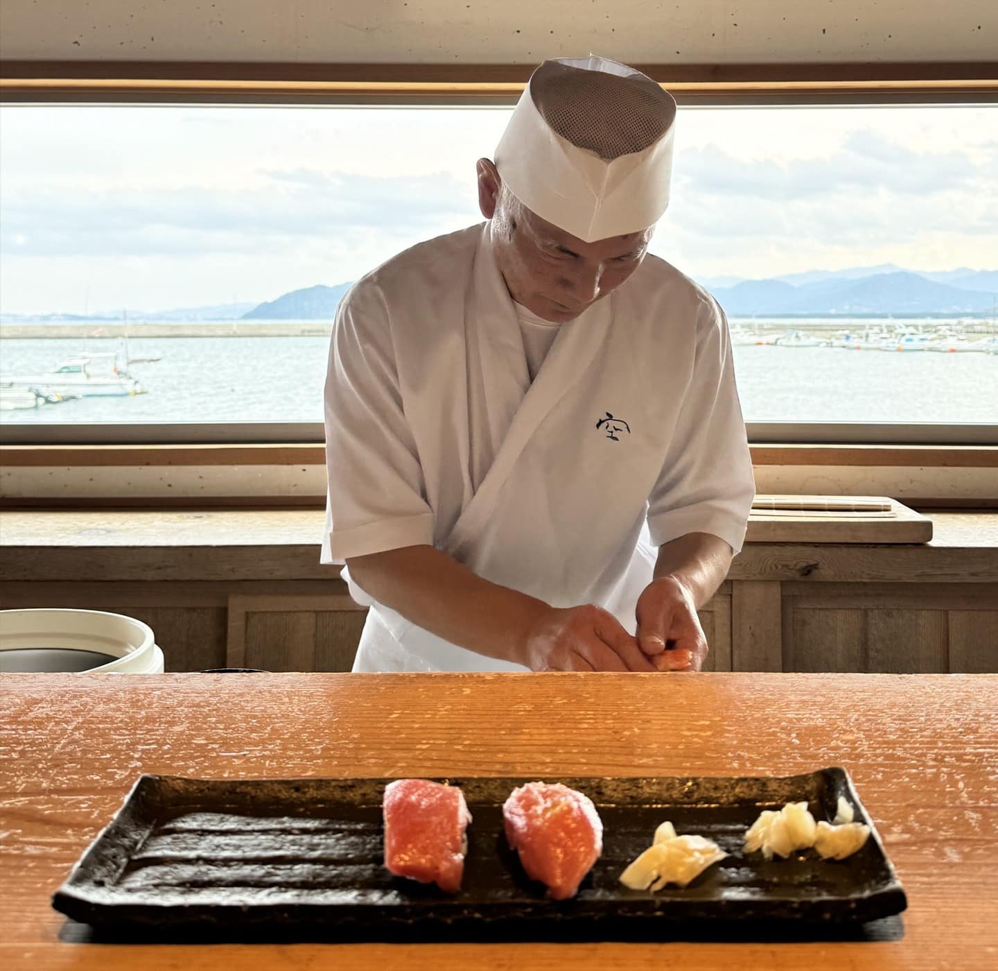 Um sushiman servindo sushis de alto nível, no fundo uma bela vista para uma baía.
