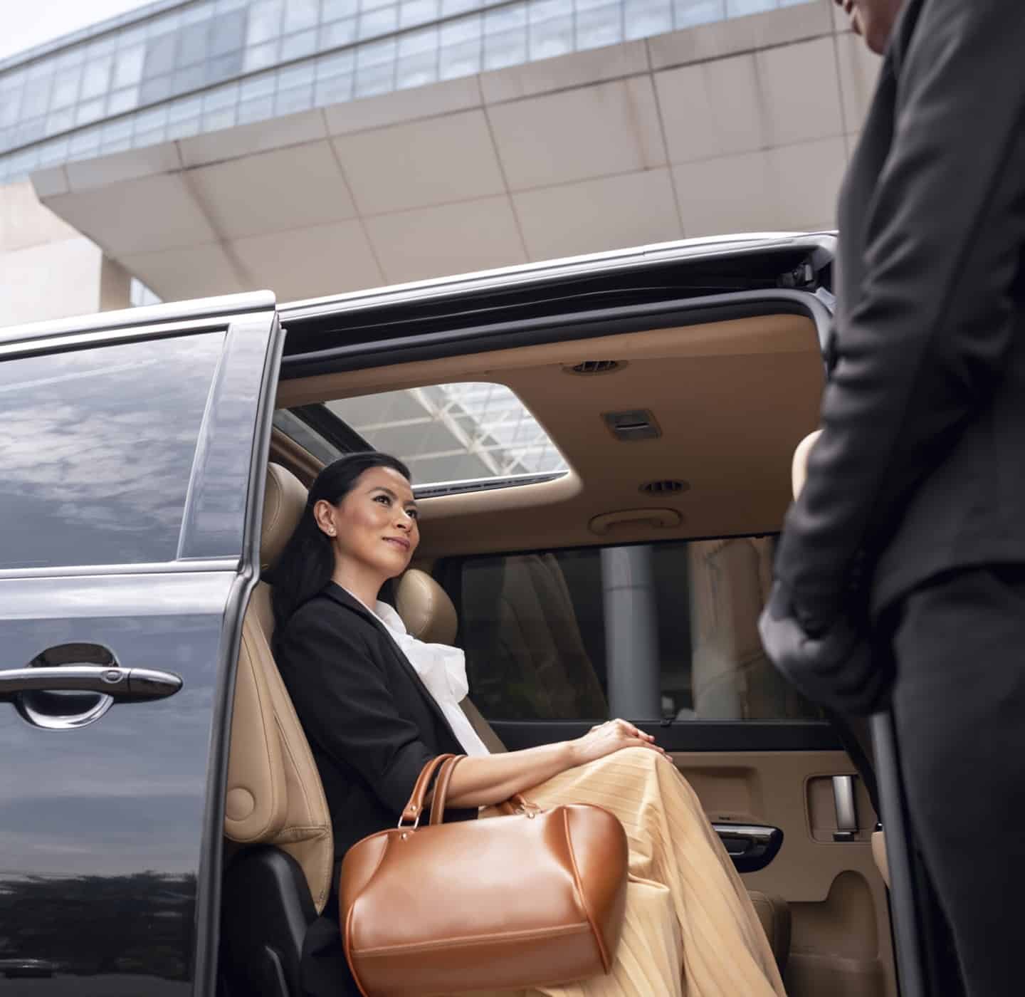 Mulher sentada num carro de luxo com a porta aberto, o motorista a aguarda do lado de fora.