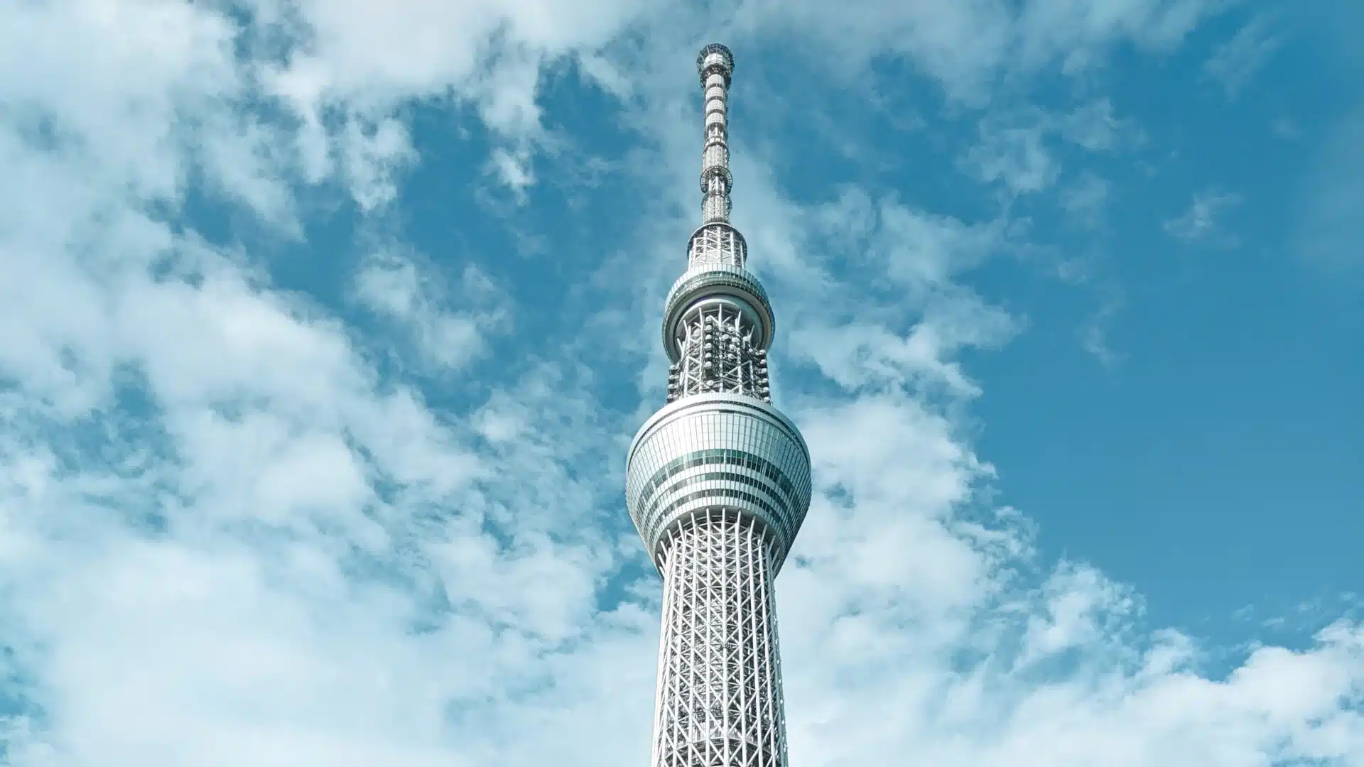 O topo da Tokyo Skytree, com estética minimalista e elegante contrastada contra o céu com algumas nuvens.