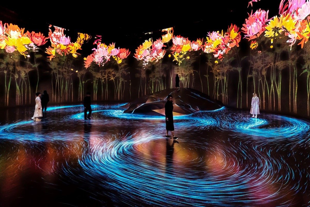 Instalação artística no museu teamLab, telas gigantes exibem flores enquanto pessoas caminham sobre um projeção, onde seus rastros de linhas azuis seguem os seus passos.